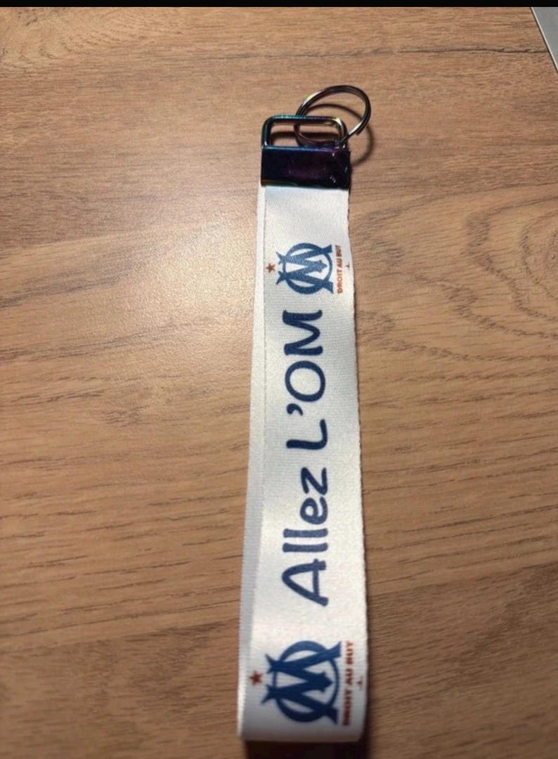 Pode incluir: Um cord&atilde;o branco com o texto "Allez L'OM" em azul, juntamente com o log&oacute;tipo da equipa. O cord&atilde;o tem um fecho met&aacute;lico e um porta-chaves. &Eacute; provavelmente um acess&oacute;rio de f&atilde;.