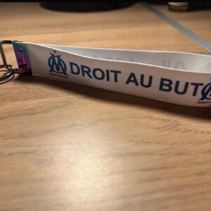 Pode incluir: Porta-chaves de tecido branco com o texto "DROIT AU BUT" em azul e um log&oacute;tipo da equipa. O porta-chaves tem um anel de metal e um fecho arco-&iacute;ris. Acess&oacute;rio para f&atilde;s de desporto.