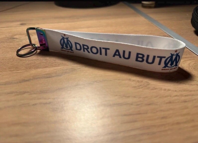 Pode incluir: Porta-chaves de tecido branco com o texto "DROIT AU BUT" em azul e um log&oacute;tipo da equipa. O porta-chaves tem um anel de metal e um fecho arco-&iacute;ris. Acess&oacute;rio para f&atilde;s de desporto.