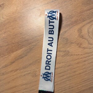 Pode incluir: Lanyard branco com o texto "DROIT AU BUT" em azul e o logotipo de uma equipa de futebol. Possui um fecho met&aacute;lico e um porta-chaves. O fundo &eacute; uma superf&iacute;cie de madeira.