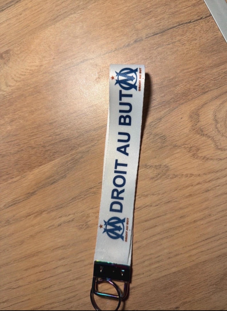 Pode incluir: Lanyard branco com o texto "DROIT AU BUT" em azul e o logotipo de uma equipa de futebol. Possui um fecho met&aacute;lico e um porta-chaves. O fundo &eacute; uma superf&iacute;cie de madeira.