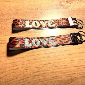 Peut inclure: Deux porte-clés avec un motif léopard orange et marron. Le mot "LOVE" est écrit en lettres argentées à paillettes sur chaque porte-clés. Chaque porte-clés a un anneau métallique.
