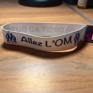 Pode incluir: Pulseira branca com o texto "Allez L'OM" em azul, juntamente com o logotipo da equipa. A pulseira tem um fecho de metal e um porta-chaves. A pulseira est&aacute; numa superf&iacute;cie de madeira.