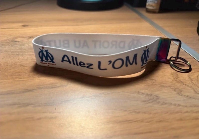 Pode incluir: Pulseira branca com o texto "Allez L'OM" em azul, juntamente com o logotipo da equipa. A pulseira tem um fecho de metal e um porta-chaves. A pulseira est&aacute; numa superf&iacute;cie de madeira.