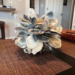 Könnte beinhalten: Eine dekorative Kugel aus zahlreichen Austernschalen, die eine Mischung aus Farben wie Weiß, Beige und Graublau aufweist. Die Schalen sind so angeordnet, dass sie einen strukturierten, geschichteten Effekt erzeugen und auf einem braunen, gewebten Tischset liegen.