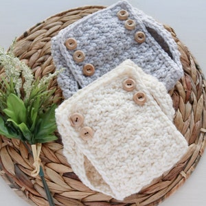 Könnte beinhalten: Zwei gehäkelte Baby-Windelüberzüge mit Holzknöpfen. Einer ist weiß und der andere grau.