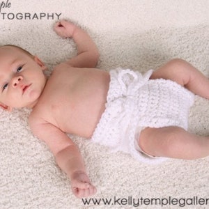 May include: A baby wearing a white crocheted diaper cover, lying on a white fluffy blanket. The baby is looking directly at the camera.