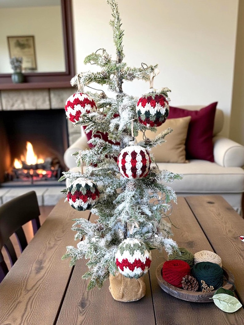May include: A small, frosted Christmas tree decorated with six handmade crochet ornaments. The ornaments feature red, white, and green chevron patterns. The tree sits on a wooden table, with a bowl of yarn and pinecones nearby.