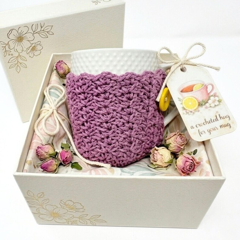 May include: A white mug with a purple crocheted cozy, presented in a decorative gift box. The box is lined with floral patterned paper and adorned with dried roses. A tag reads "a crocheted hug for your mug."