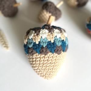May include: Crocheted acorn with a light tan base and a cap featuring a pattern of white, tan, teal, and brown. The acorn has a wooden stem. Several other crocheted acorns are visible in the background.
