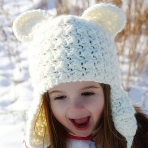 May include: A white crocheted hat with bear ears and ear flaps. The hat is worn by a child with long brown hair. The child is smiling and looking at the camera.