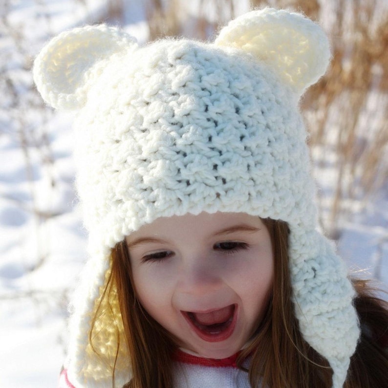 May include: A white crocheted hat with bear ears and ear flaps. The hat is worn by a child with long brown hair. The child is smiling and looking at the camera.