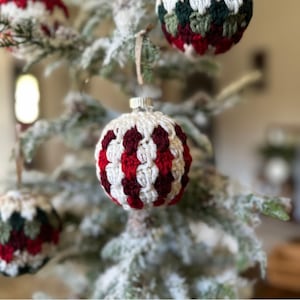 May include: Close-up of a Christmas tree adorned with handmade crochet ornaments. The ornaments feature a white base with red and burgundy accents. The tree branches are dusted with artificial snow, creating a festive holiday scene.