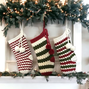 May include: Three crocheted Christmas stockings hanging on a fireplace mantel. The stockings are red, white and green with a striped pattern. Each stocking has a white pom-pom at the top.