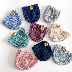 May include: A collection of handmade crochet coasters in various colors, including blue, pink, and beige. Each coaster features a scalloped edge and a wooden button detail. The coasters are arranged on a white surface.