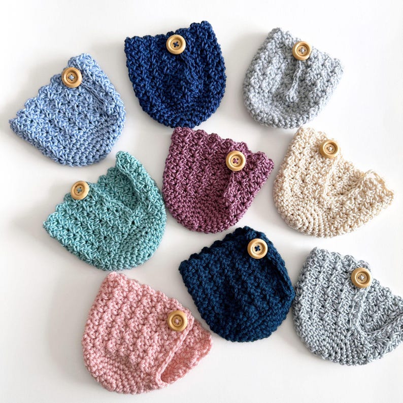 May include: A collection of handmade crochet coasters in various colors, including blue, pink, and beige. Each coaster features a scalloped edge and a wooden button detail. The coasters are arranged on a white surface.