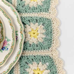 May include: A crocheted table runner with a scalloped edge. The runner is made of white and green yarn and features a repeating flower pattern.