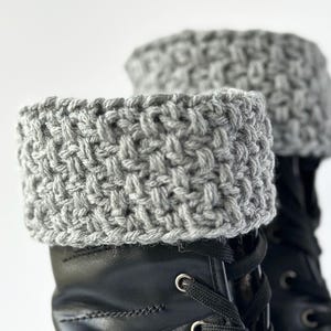 Puede incluir: Polainas de bota de crochet gris, hechas con un patrón de puntada texturizada, se muestran en un par de botas de cuero negro con cordones.