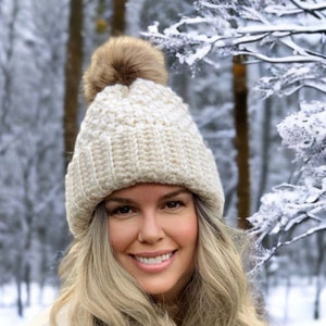 a women wearing a cream thick crochet hat with a rolled brim and knit look texture with pompom snow in the background