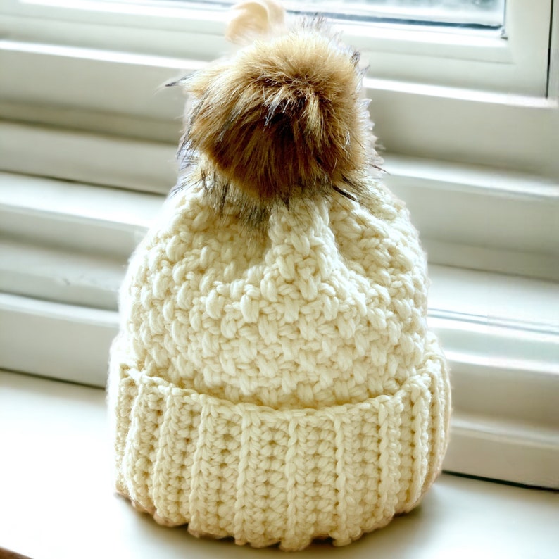 a cream thick crochet hat with a rolled brim and knit look texture with pompom on a window sill