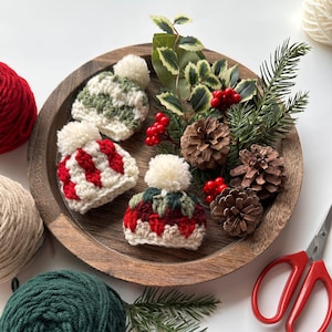 May include: Three miniature crocheted hats with white pom-poms are arranged on a wooden tray. One hat features red and white stripes, another has green and white patterns, and the third has red, green, and white designs. The tray is decorated with pinecones, berries, and greenery.
