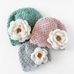 May include: Three crocheted hats with flower embellishments. The hats are in shades of green, pink, and gray. Each hat has a white crocheted flower with a brown button in the center.