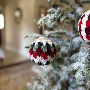 May include: A handmade crochet Christmas ornament with a festive color scheme. The sphere-shaped ornament features alternating rows of red, green, cream, and dark green yarn. It hangs from a natural twine loop, ready for display on a holiday tree.