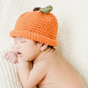 May include: A bright orange crochet hat featuring a textured, rounded design with a small brown stem and two green leaves attached to the top. The hat is crafted with a thick yarn, creating a visible knit pattern that covers the crown of the head.