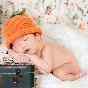 May include: A newborn baby wearing an orange crocheted pumpkin hat sleeps on a white fluffy blanket. The baby is lying on its side with its head resting on its arm.