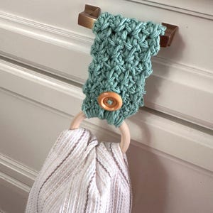 May include: A sage green crocheted towel holder featuring a light wood ring and a single wooden button. The holder is looped over a metallic cabinet handle and secures a white waffle-weave kitchen towel with thin brown vertical stripes.