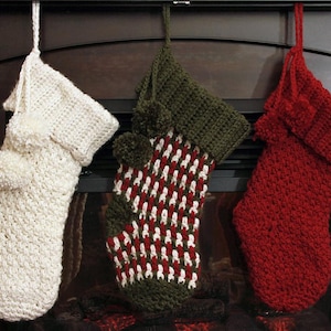 Puede incluir: Tres calcetines de Navidad tejidos a ganchillo colgados en fila. El calcetín de la izquierda es de color crema sólido, el del medio presenta un patrón tejido en rojo, blanco y verde oliva, y el de la derecha es rojo sólido. Cada uno tiene un puño y pompones.