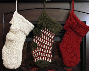 Könnte beinhalten: Drei gehäkelte Weihnachtsstrümpfe hängen in einer Reihe. Der linke Strumpf ist cremefarben, der mittlere hat ein gewebtes Muster in Rot, Weiß und Olivgrün, und der rechte ist einfarbig rot. Jeder Strumpf hat einen Umschlag und Bommeln.