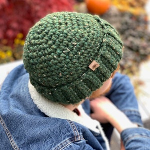 Può includere: Un berretto all'uncinetto verde bosco scuro con una finitura in filato testurizzato e maculato. Il cappello presenta un bordo a coste e una piccola etichetta quadrata in similpelle marrone sul lato, con il disegno di un uccellino su un ramo.