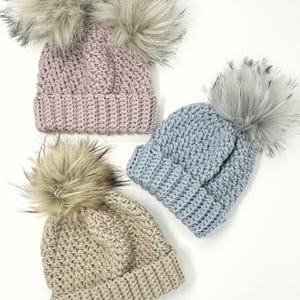 May include: Three crocheted hats with faux fur pom-poms. The hats are in pink, blue, and beige.