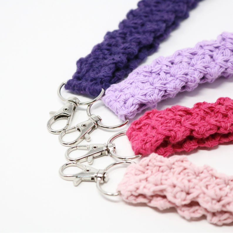May include: Close-up of four crocheted lanyards in shades of purple, lavender, pink, and light pink. Each lanyard has a silver-colored metal clasp. The lanyards are made of a textured, woven material.