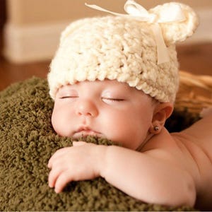 May include: A sleeping baby wearing a white crocheted hat with bear ears and a bow. The hat has a cream color and is made of soft yarn. The baby is lying on a green fuzzy blanket.
