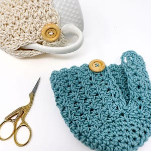 May include: A close-up shot of two crocheted cup cozies, one cream-colored and the other teal, each with a wooden button closure. A pair of gold-colored scissors is also in the frame. The cozies are designed to fit around a mug.