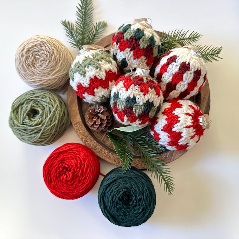 May include: A wooden tray holds six crocheted Christmas ornaments in red, white, and green patterns. Several balls of yarn in beige, green, red, and dark green are arranged around the tray, along with pine needles and a pine cone. Festive holiday decor.