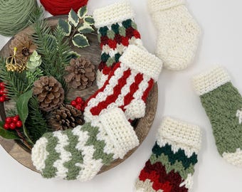 May include: A collection of miniature, hand-crocheted Christmas stockings in various colors and patterns. The stockings feature red, green, and white designs, with a cream-colored cuff. The arrangement includes yarn balls, pinecones, and greenery, creating a festive holiday theme.