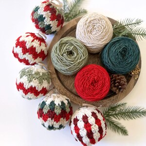 May include: A wooden tray holds several crochet Christmas ornaments in red, white, and green. Also on the tray are balls of yarn in various colors, including cream, green, and red. Pine needles and a pine cone add to the festive theme.