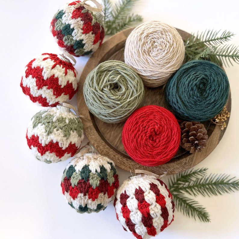 May include: A wooden tray holds several crochet Christmas ornaments in red, white, and green. Also on the tray are balls of yarn in various colors, including cream, green, and red. Pine needles and a pine cone add to the festive theme.