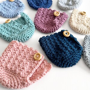 May include: A collection of handmade crochet cozies in various colors, including pink, teal, and navy blue. Each cozy features a scalloped edge and is secured with a wooden button. These items are ideal for protecting cups and mugs.