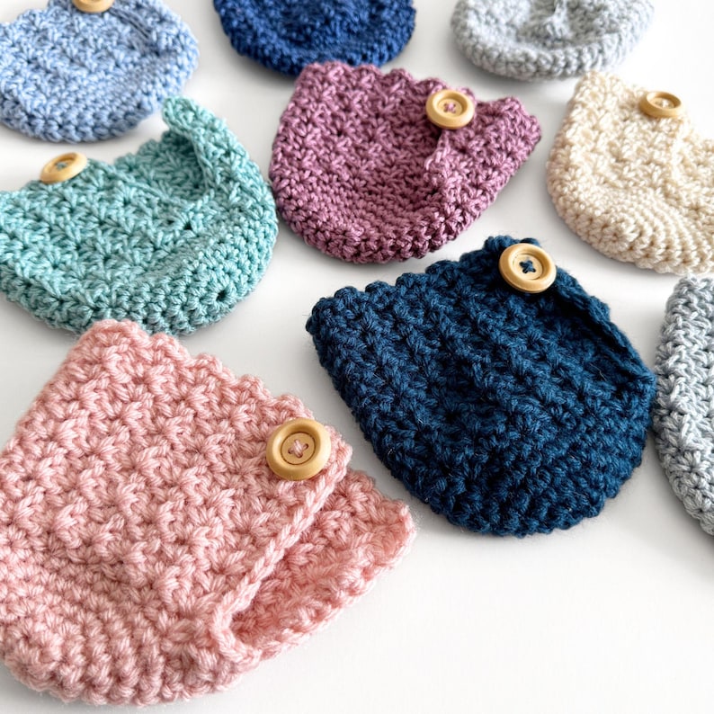 May include: A collection of handmade crochet cozies in various colors, including pink, teal, and navy blue. Each cozy features a scalloped edge and is secured with a wooden button. These items are ideal for protecting cups and mugs.