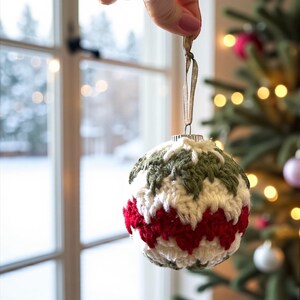May include: A handmade Christmas ornament with a crocheted design. The ornament features a pattern of red, white, and green yarn. It is suspended by a gold ribbon, and is displayed in front of a Christmas tree.