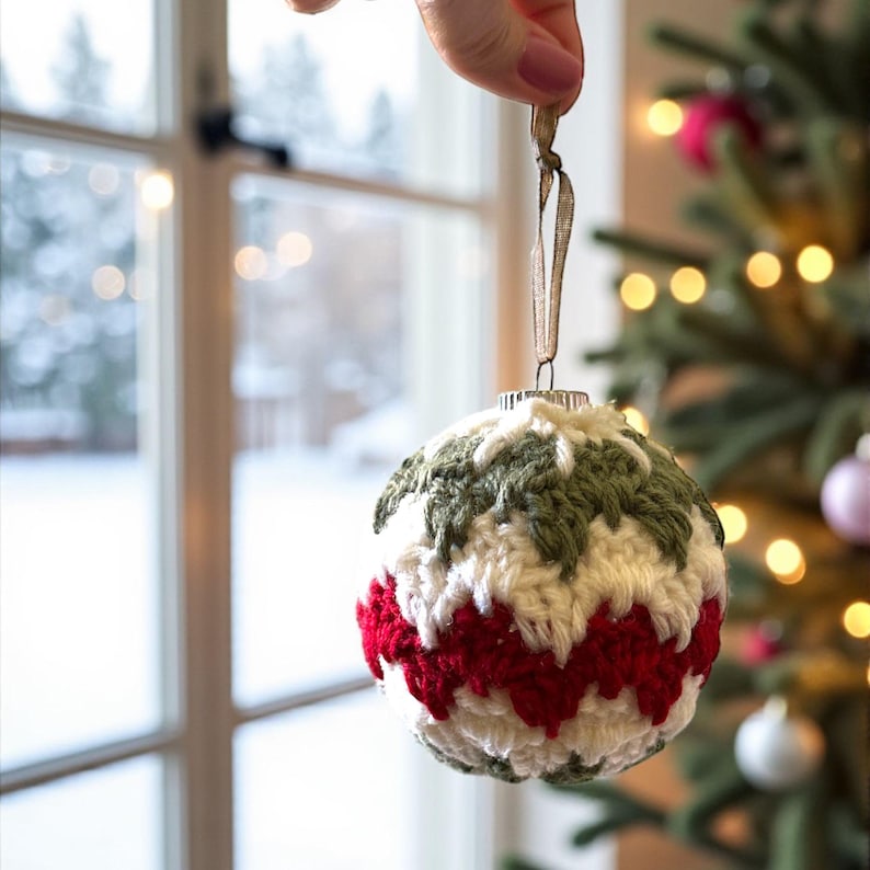 May include: A handmade Christmas ornament with a crocheted design. The ornament features a pattern of red, white, and green yarn. It is suspended by a gold ribbon, and is displayed in front of a Christmas tree.