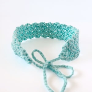 Könnte beinhalten: Ein hellblaues gehäkeltes Stirnband mit langen Bändern. Das Stirnband ist aus weichem, strukturiertem Garn.