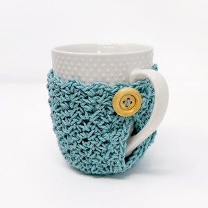 May include: A white ceramic mug with a textured surface is wrapped in a teal crocheted cozy. The cozy is secured with a wooden button. The mug's handle is visible, and the background is white.