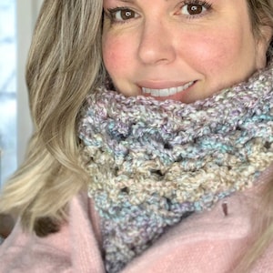 May include: A close-up of a woman wearing a multi-coloured crocheted neck warmer. The neck warmer is made with a variety of shades of purple, blue, and beige yarn.