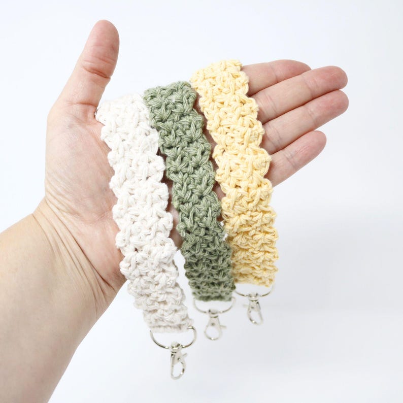 May include: Three crocheted wrist straps in cream, sage green, and pale yellow. Each strap has a silver-colored clasp. The straps are made of a textured, woven material. The wrist straps are held in a hand against a white background.