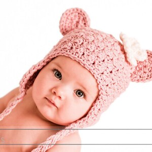 May include: A baby wearing a pink crocheted hat with bear ears and a white flower accent. The hat has long ties that can be tied under the chin.